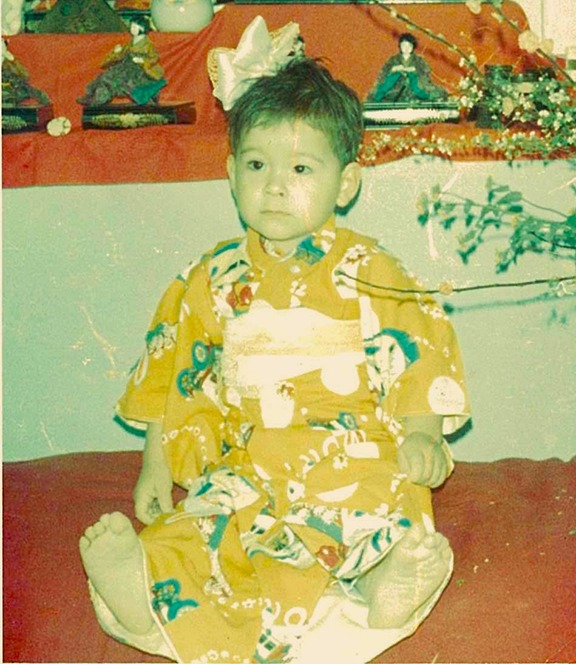 Adorable picture of baby Val in a gold kimono that has colorful designs. She also has a big bow on her head. There are Japanese decorative pieces in the background. She looks uncertain about posing for this picture, and is likely wondering when her next snack is.