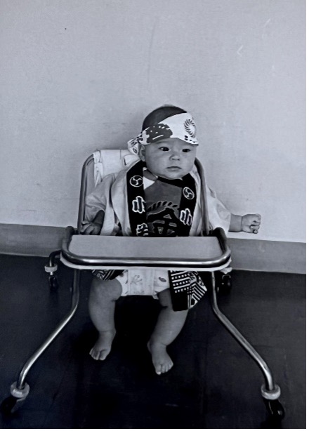 Baby Val in a walker with a band tied around her head that has Japanese designs on it. She is wearing a yukata. She is likely waiting for someone to put some snacks on the tray of her walker.