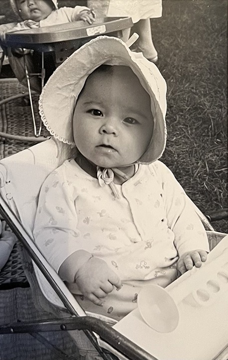 Baby Val in a stroller wearing a baby bonnet. She has a questioning look, probably wondering about a snack.